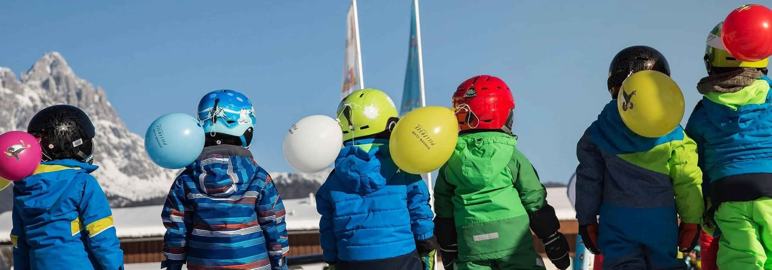 Kinder im Skianzug mit Helmen und Luftballons vor verschneiten Bergen der